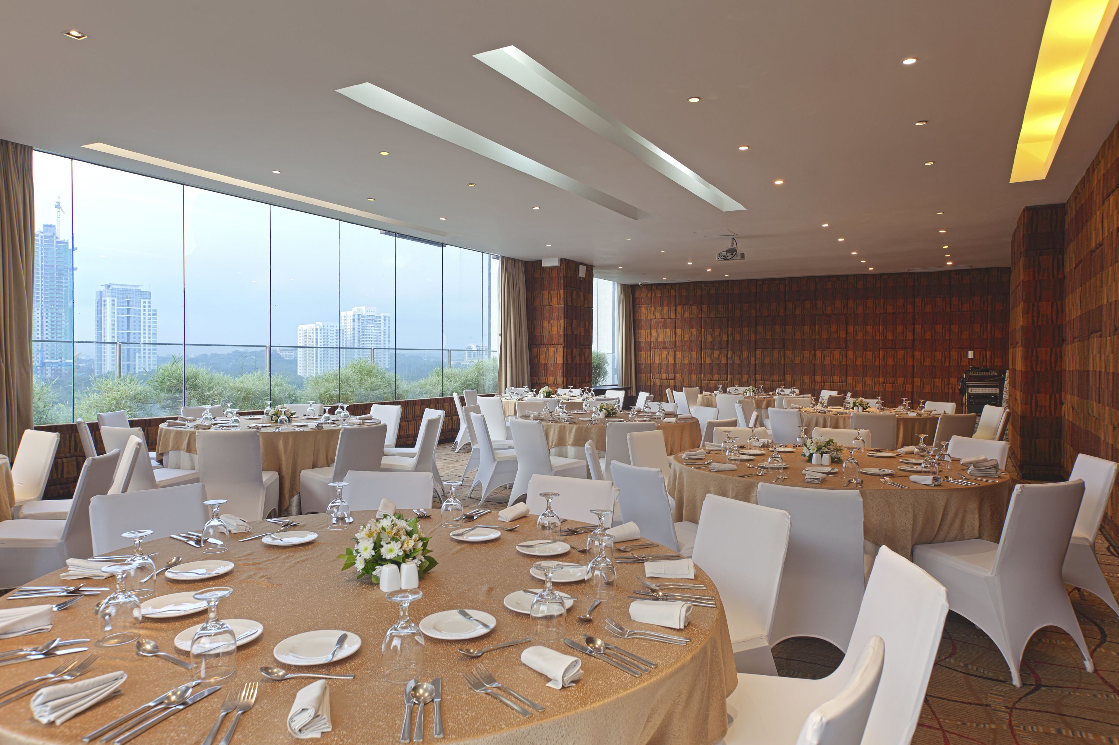A spacious banquet hall with multiple round tables set for dining, large windows offering a city view, and a modern ceiling design.
