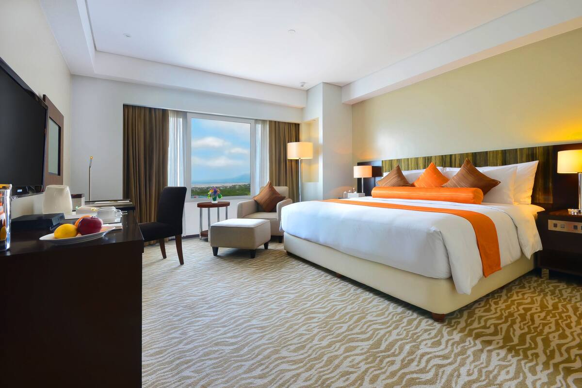 A hotel room with a large bed, a desk with a flat-screen TV, a chair, a small table, and a view of the outdoors.