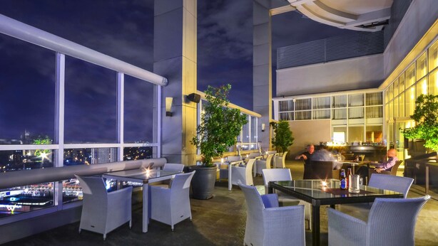 An outdoor dining area with tables and chairs, large windows, and a view of the city at night. - Acacia Hotel Manila (Muntinlupa)
