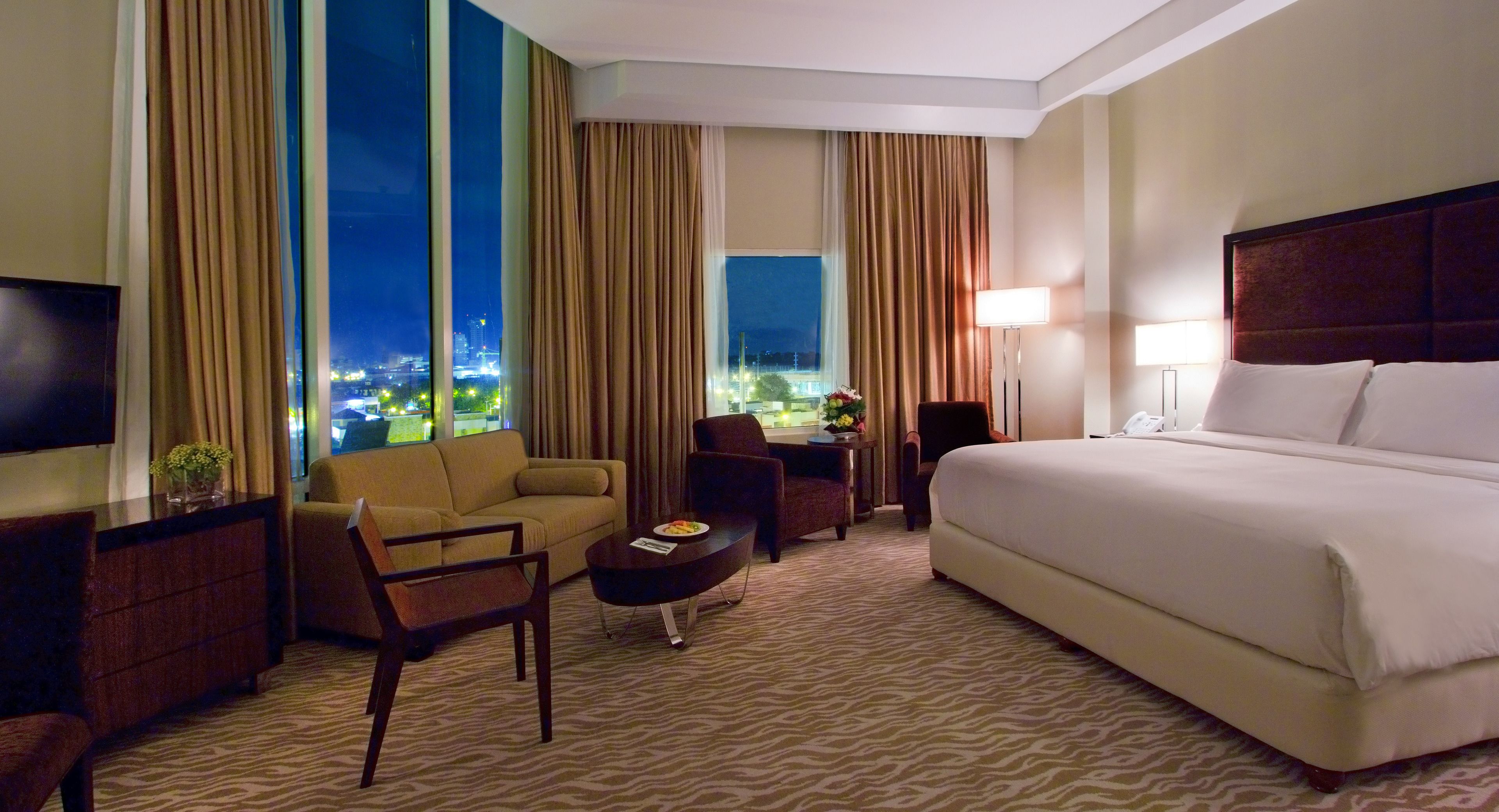 Habitación de hotel con una cama grande, un sofá, un escritorio y una silla. Hay un televisor a la izquierda y una ventana con cortinas.