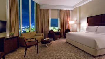 Habitación de hotel con una cama grande, un sofá, un escritorio y una silla. Hay un televisor a la izquierda y una ventana con cortinas.