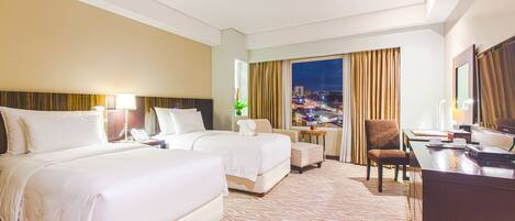 A hotel room with two beds, a desk, a chair, a TV, and a large window with a city view.