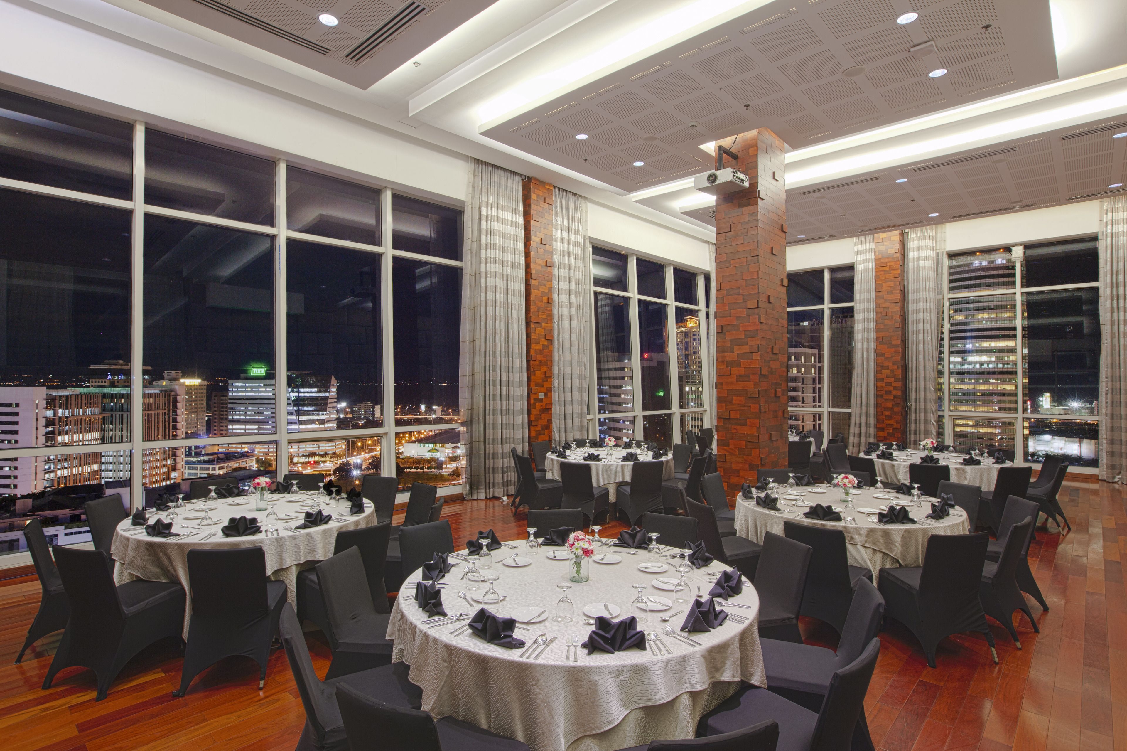 A restaurant with round tables set for dining, large windows with curtains, and a city view.