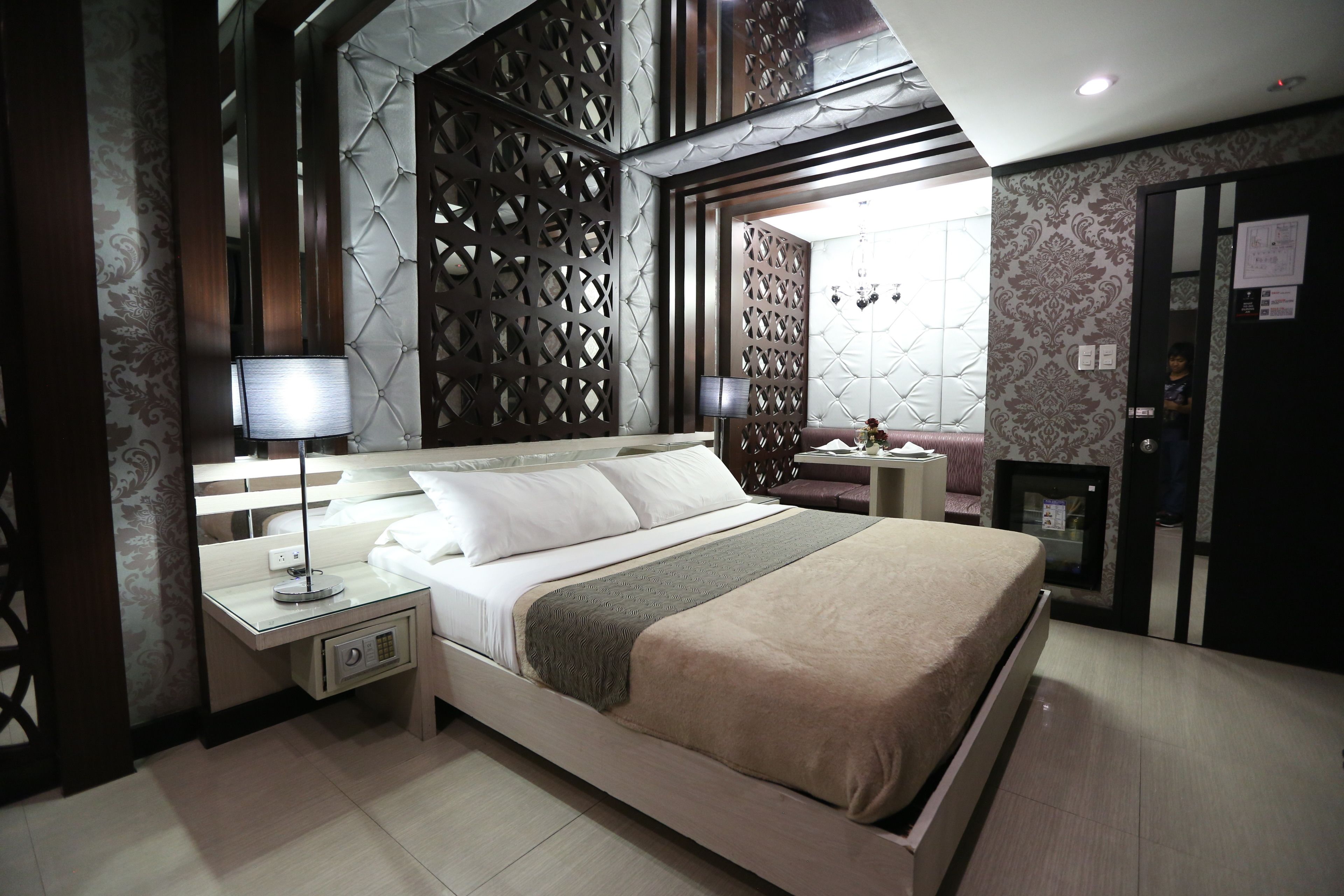 A modern bedroom with a large bed, bedside tables with lamps, a decorative wall panel, and a small seating area with a chair and table.