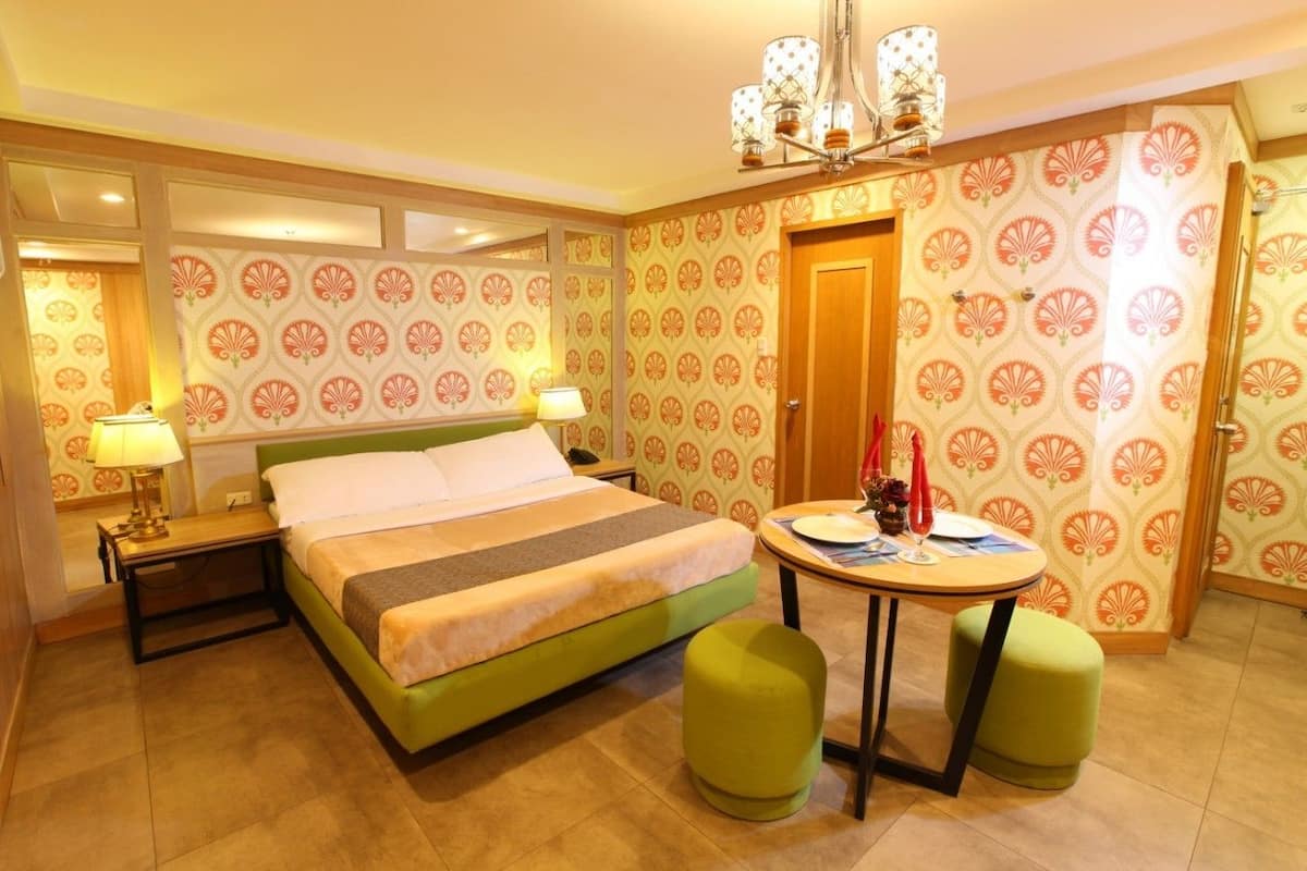 Une chambre d’hôtel avec un lit, une table ronde, deux poufs verts, un lustre et un papier peint à motifs.