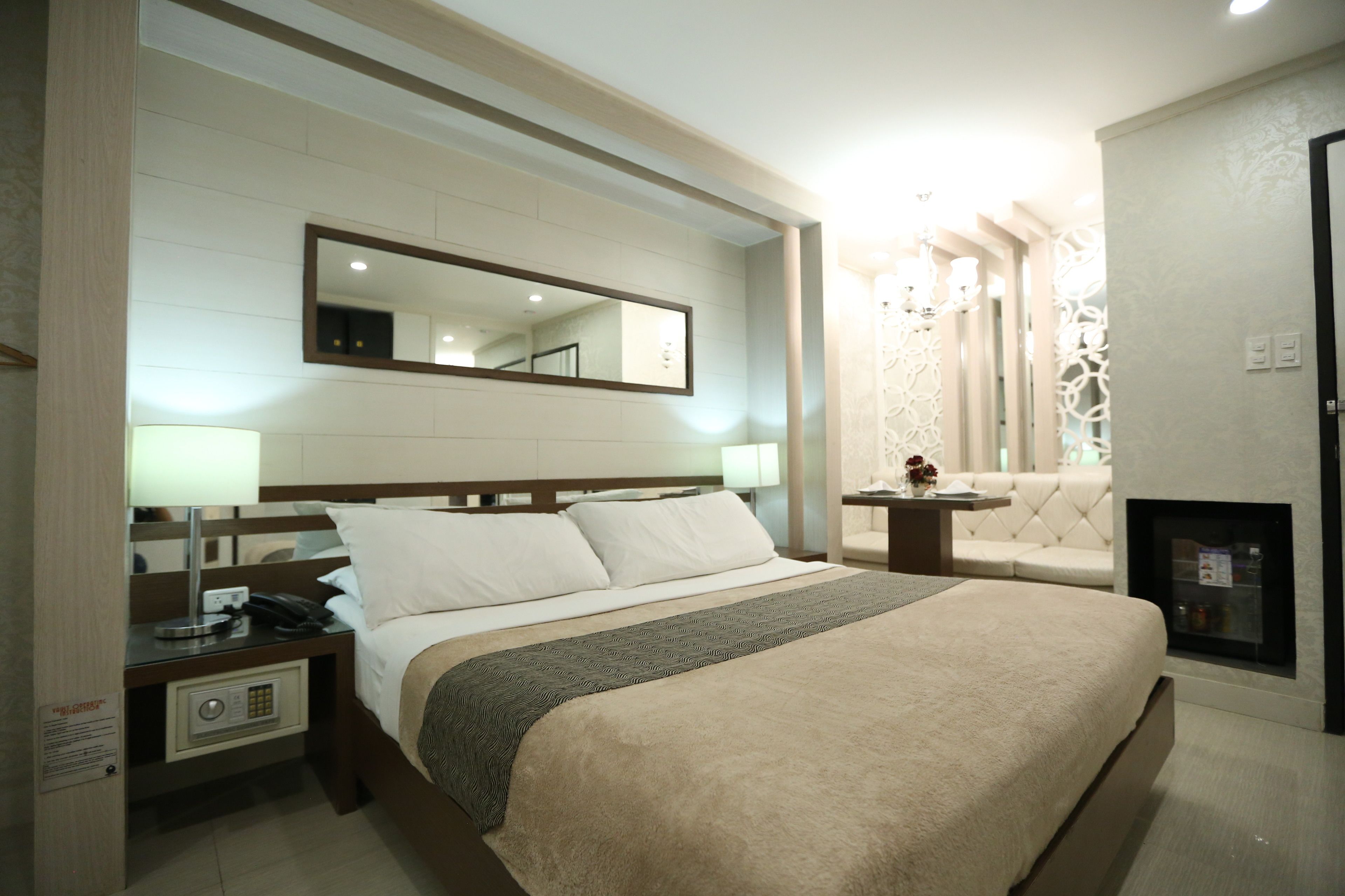 A hotel room with a large bed, a bedside table with a lamp, a nightstand with a phone, a wall-mounted mirror, and a fireplace.