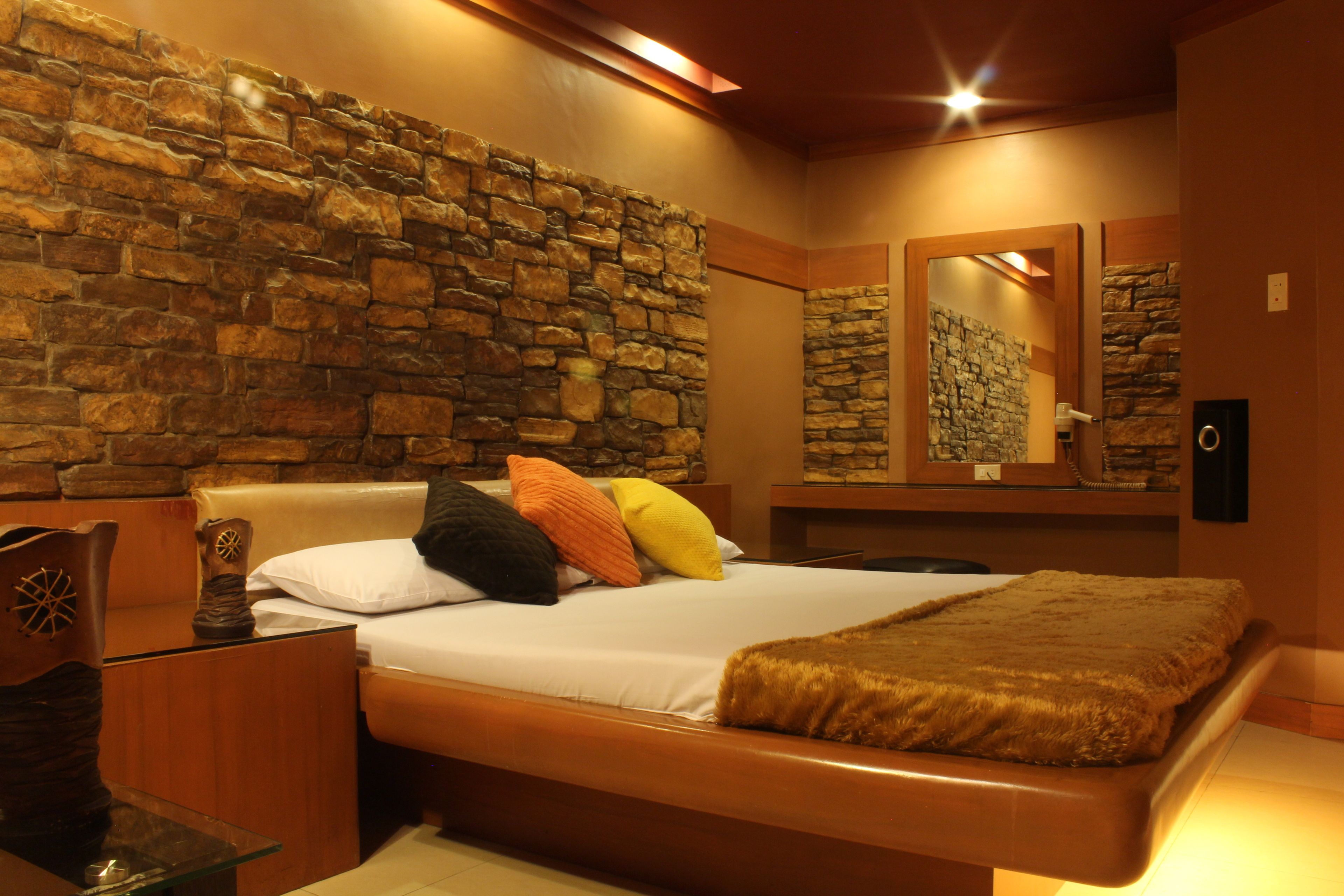 A bedroom with a stone wall, a bed with multiple pillows, a wooden headboard, a bedside table, and a mirror on the wall.