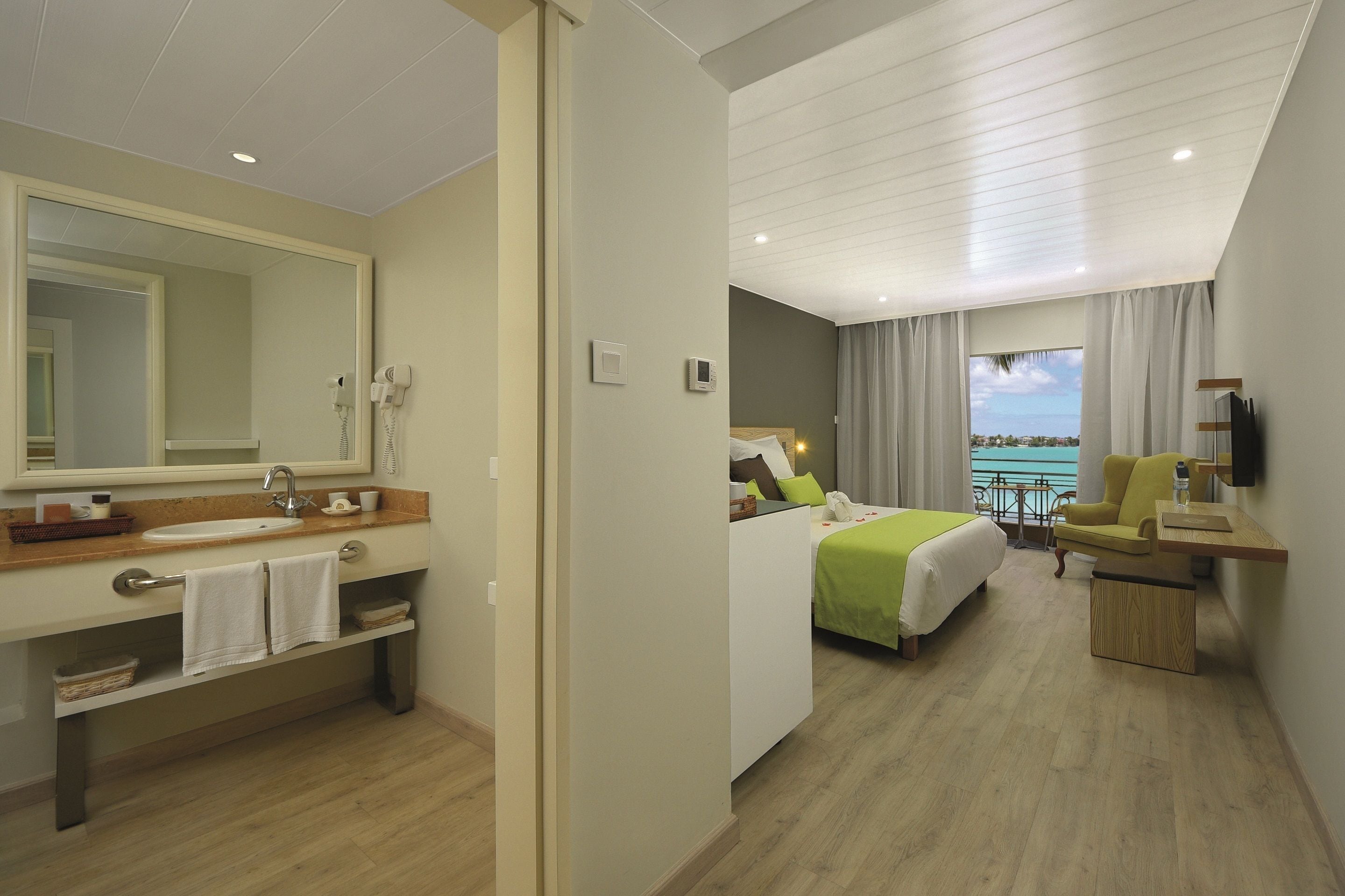 A hotel room with a large mirror, a sink, a bed with green bedding, a chair, and a desk.