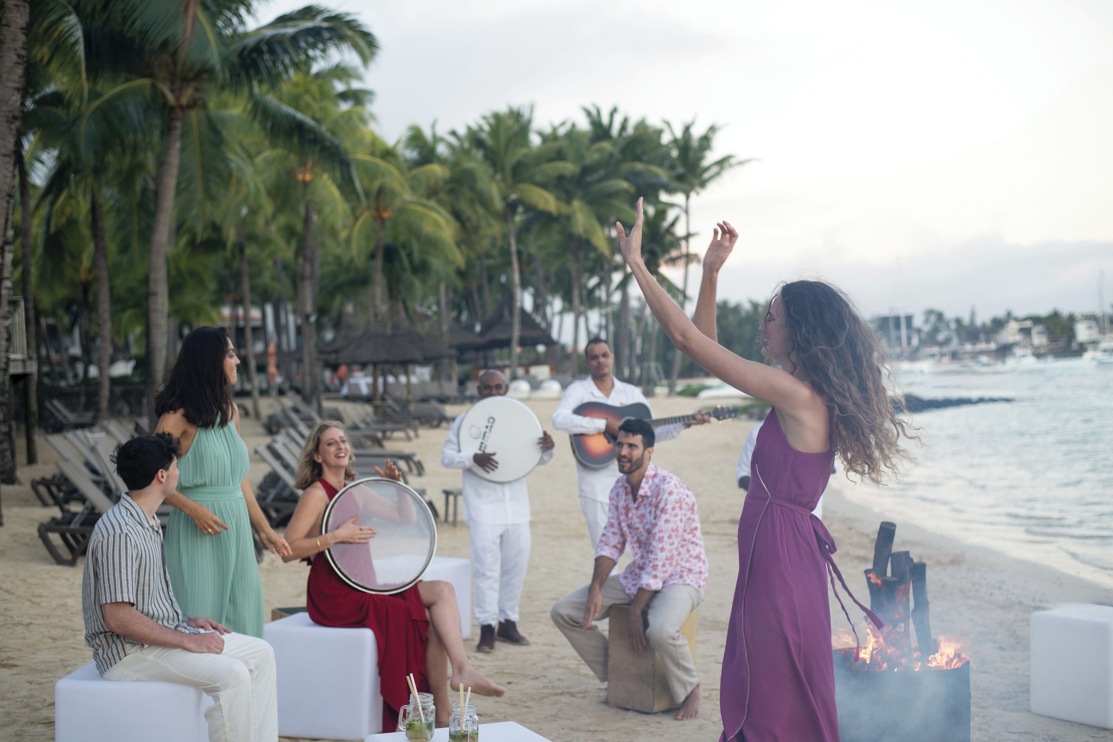 A beachside gathering with people seated and standing, palm trees, and a fire pit in the background.