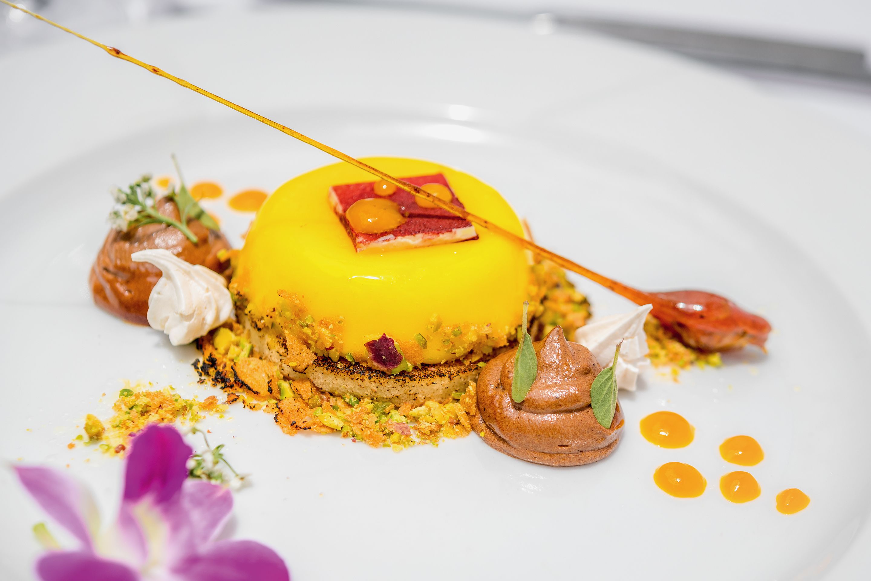 Un dessert surmonté d’une sorte de dôme jaune, décoré d’un carré rouge et d’une feuille verte, présenté sur une assiette blanche parsemée de points jaunes et agrémenté d’une fleur violette.