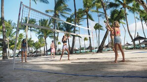 Ein Beachvolleyballspiel mit Spielern und Netz, umgeben von Palmen und Strandsonnenschirmen.