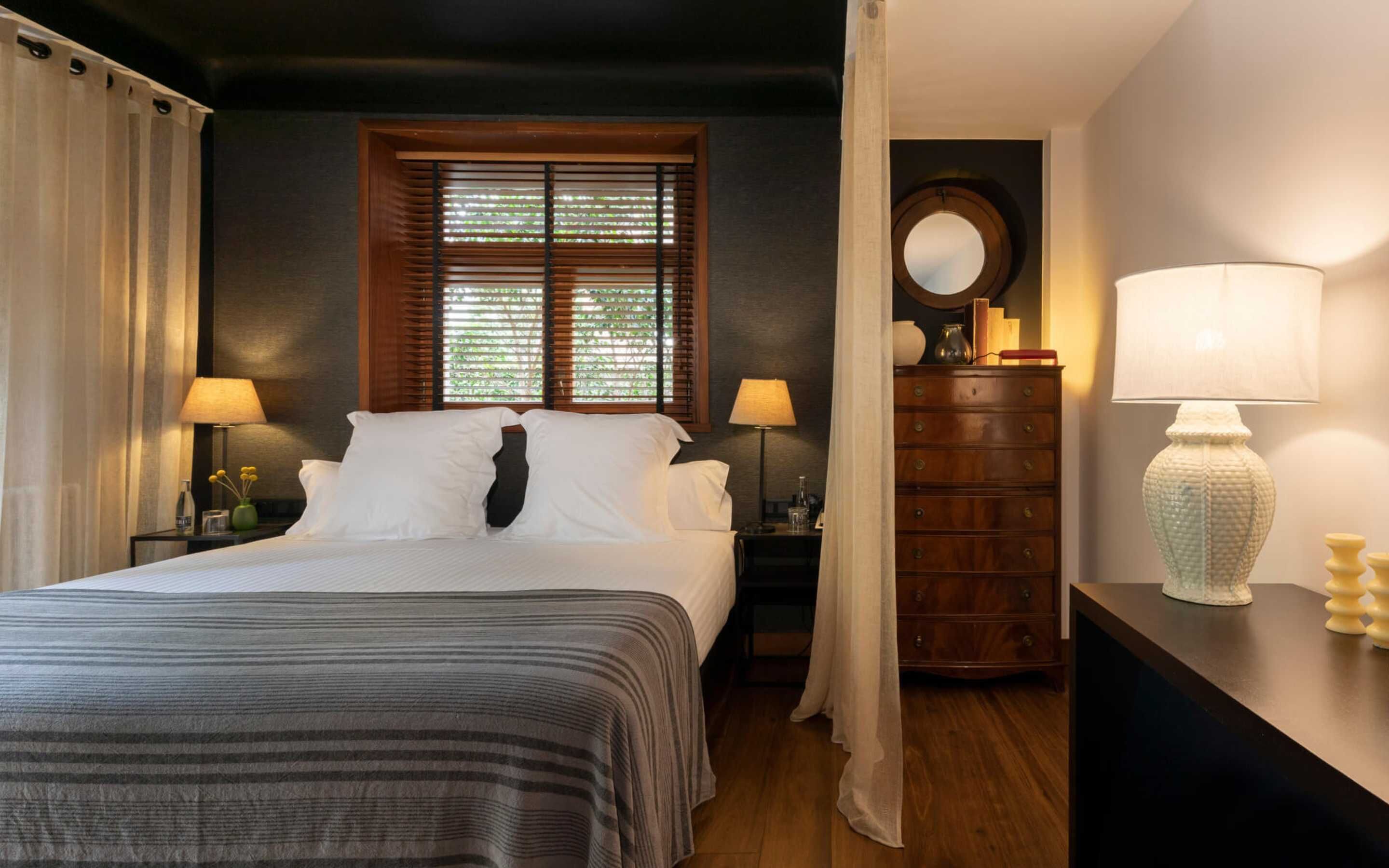 A hotel room with a bed, a dresser, a nightstand, a lamp, and a window with blinds.