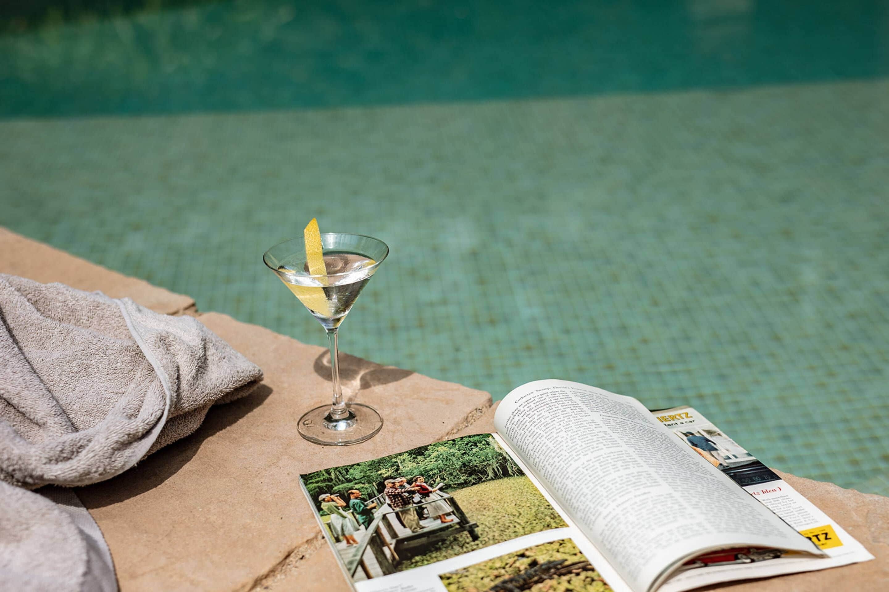 Un magazine, un verre à martini orné d’une tranche de citron et une serviette près d’une piscine.