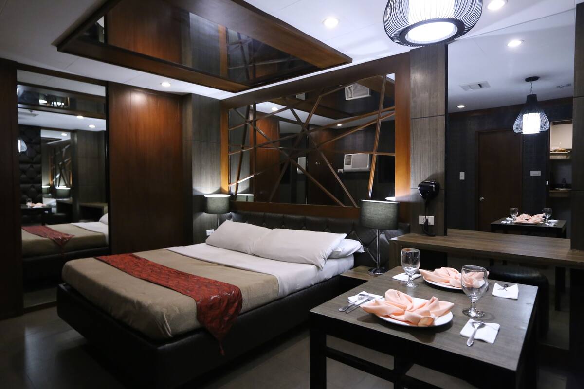 A modern hotel room with a large bed, a dining table with plates and glasses, and a unique geometric ceiling design.