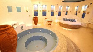 A modern spa room with a circular hot tub, a round bed with pillows, a small table with chairs, and a view of the sea through windows. - Victoria Court Malabon (Malabon)