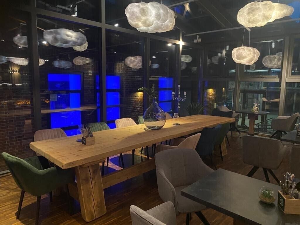 Uma área de refeições moderna com mesa e cadeiras de madeira, janelas amplas com iluminação azul e luminárias em formato de nuvem penduradas.