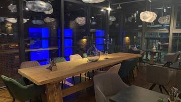 Ein moderner Essbereich mit Holztisch und -stühlen, großen Fenstern mit blauer Beleuchtung und hängenden, wolkenförmigen Lampen.