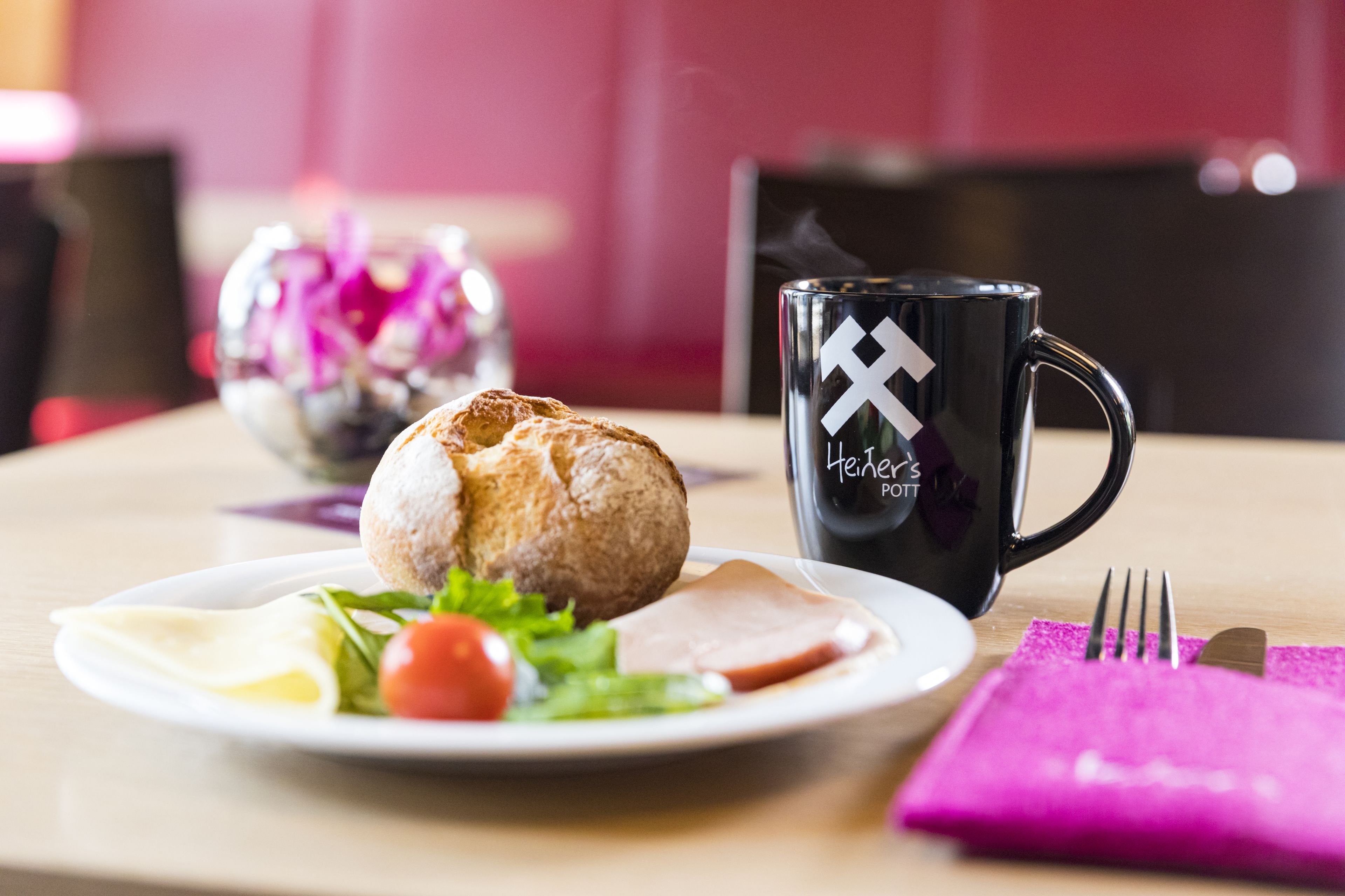 A plate with a croissant, a slice of ham, a tomato, and some greens on a white plate. There's a black mug with a logo and steam rising from it.