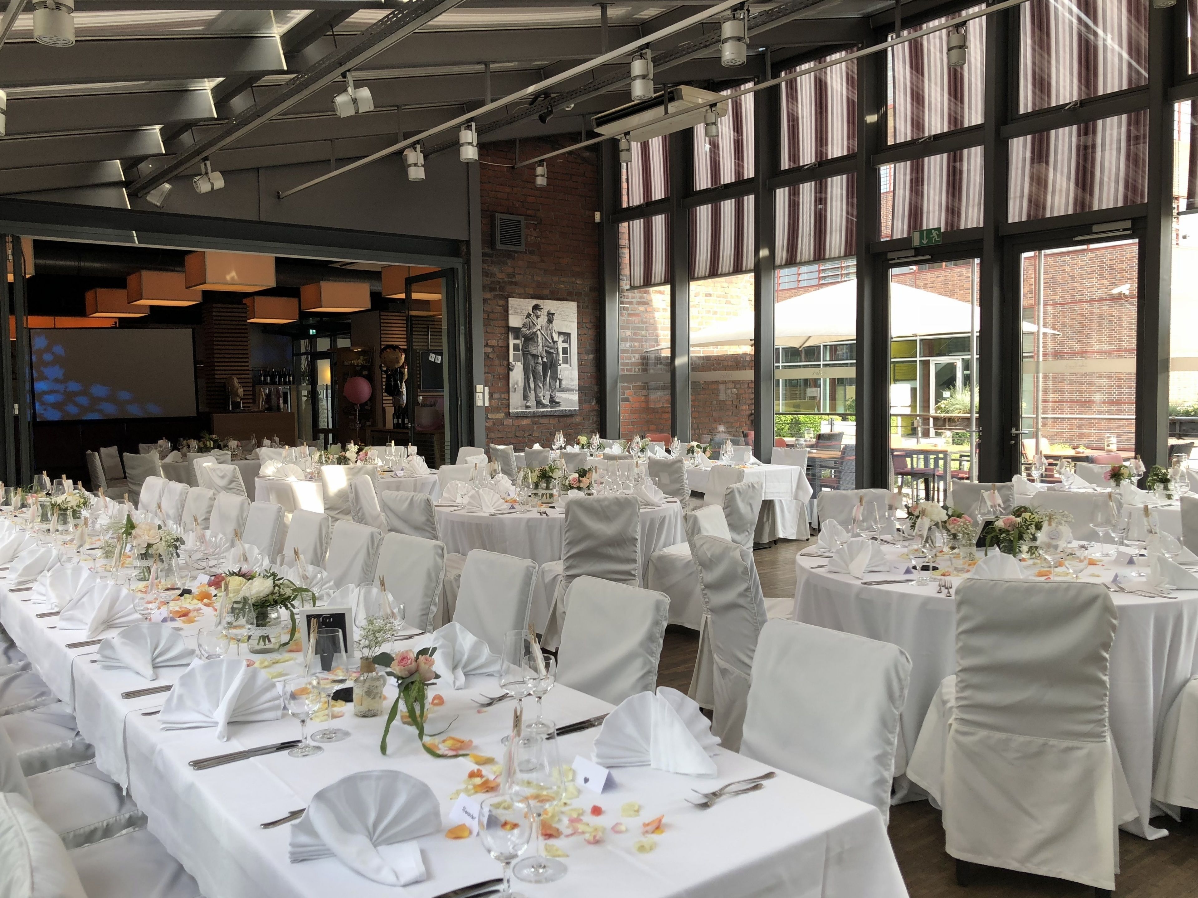 Une salle de banquet avec de nombreuses tables rondes dressées pour un événement formel, agrémentée de nappes blanches, de chaises et de compositions florales.