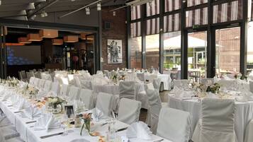 Une salle de banquet avec de nombreuses tables rondes dressées pour un événement formel, agrémentée de nappes blanches, de chaises et de compositions florales.