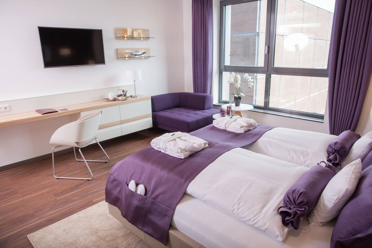 A modern hotel room with a large bed, a desk, a chair, and a TV on the wall.