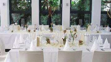 A formal dining room with white tablecloths, white chairs, and a view of greenery through large windows.