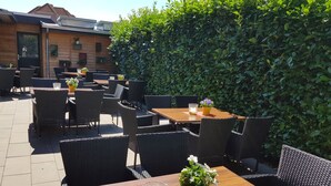 Eine Terrasse im Freien mit Holztischen und Korbstühlen, umgeben von einer Hecke.