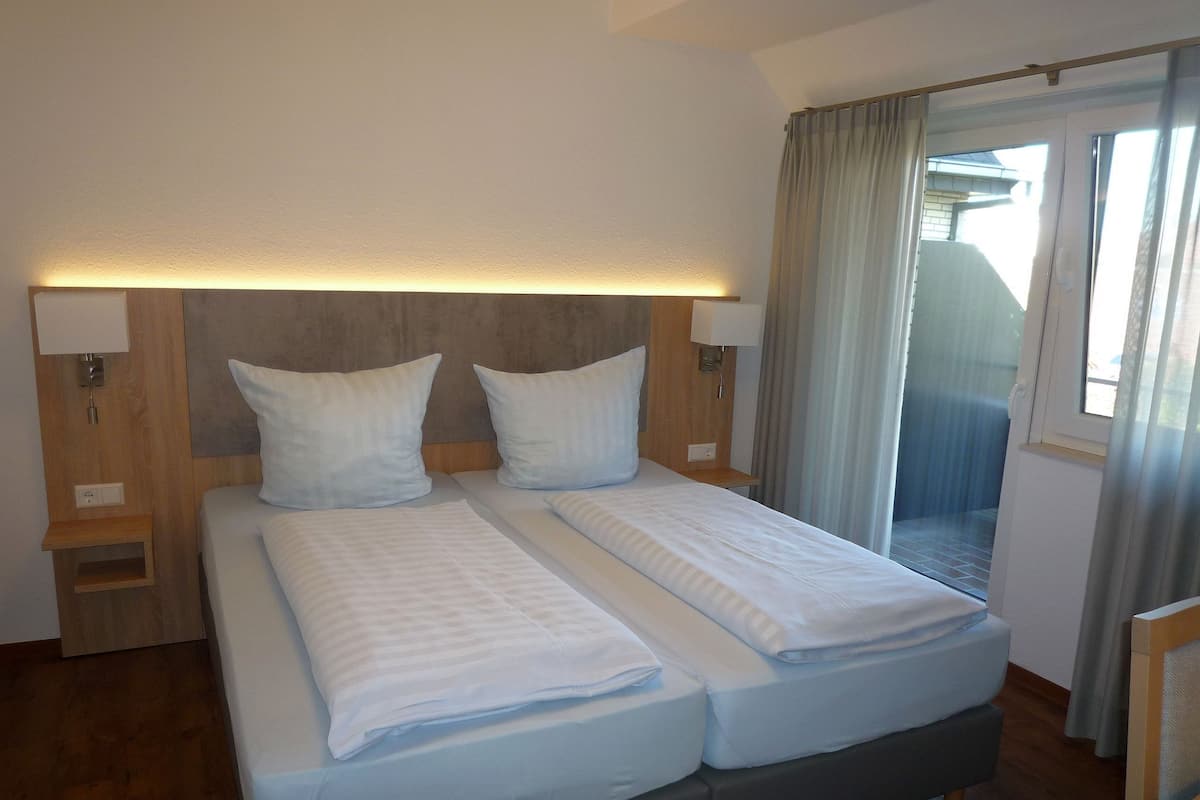 A double bed with two pillows, a wooden headboard, bedside lamps, and a view of a balcony through a glass door.