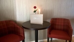 Ein Zimmer mit zwei roten Polsterstühlen, einem kleinen runden Tisch mit einer weißen Schachtel mit dem Aufdruck "Verscholten" und einer roten Rose darauf, sowie einer strukturierten Wand im Hintergrund.