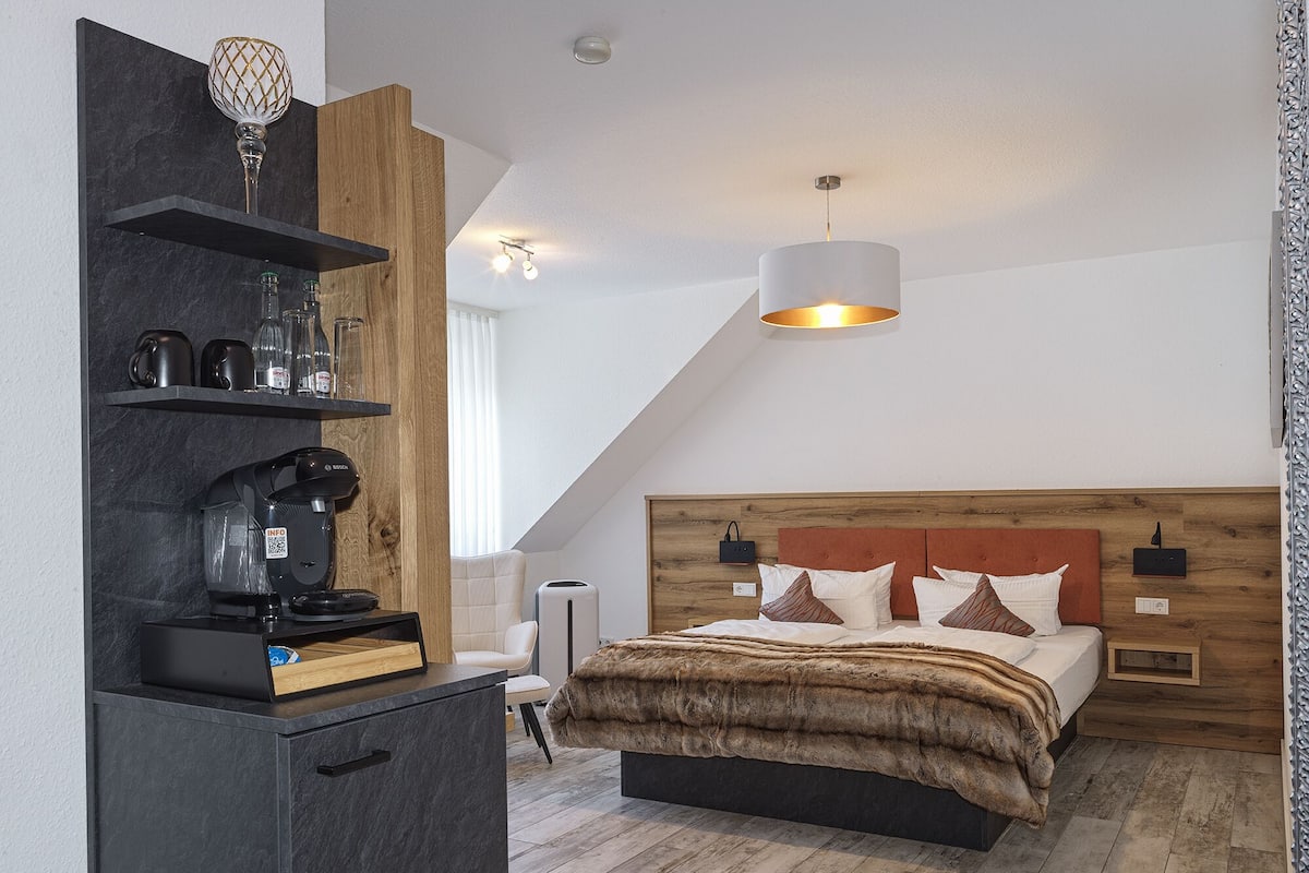 Une chambre moderne avec une tête de lit en bois, un lit recouvert d’un plaid en fausse fourrure marron, une machine à café et une étagère avec de la verrerie.