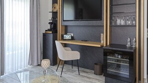 Una sala moderna con un televisor de pantalla plana montado en una pared oscura, un escritorio de madera con una silla, una cafetera y un aparador de vidrio con copas de vino.