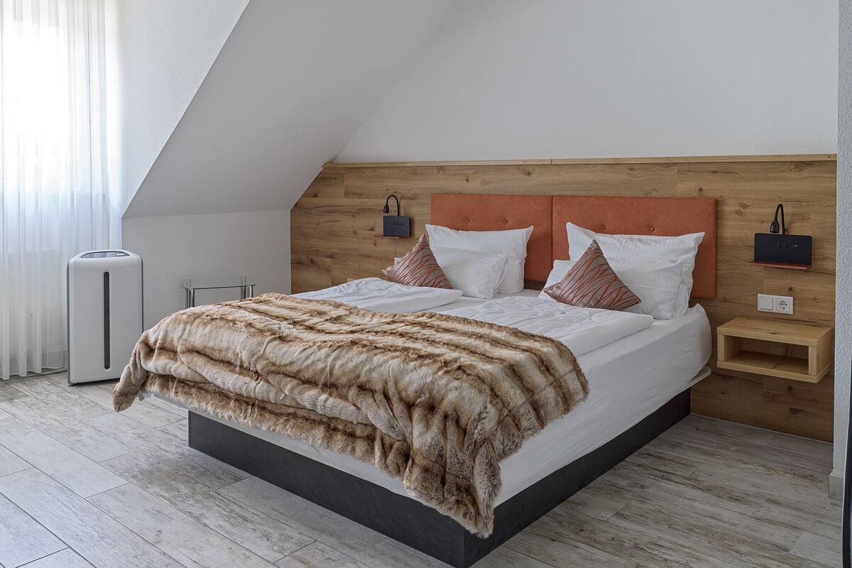 Une chambre moderne avec un plafond en pente, une tête de lit en bois, un grand lit avec une literie blanche et des coussins à motifs, des tables de chevet avec des lampes et un radiateur.