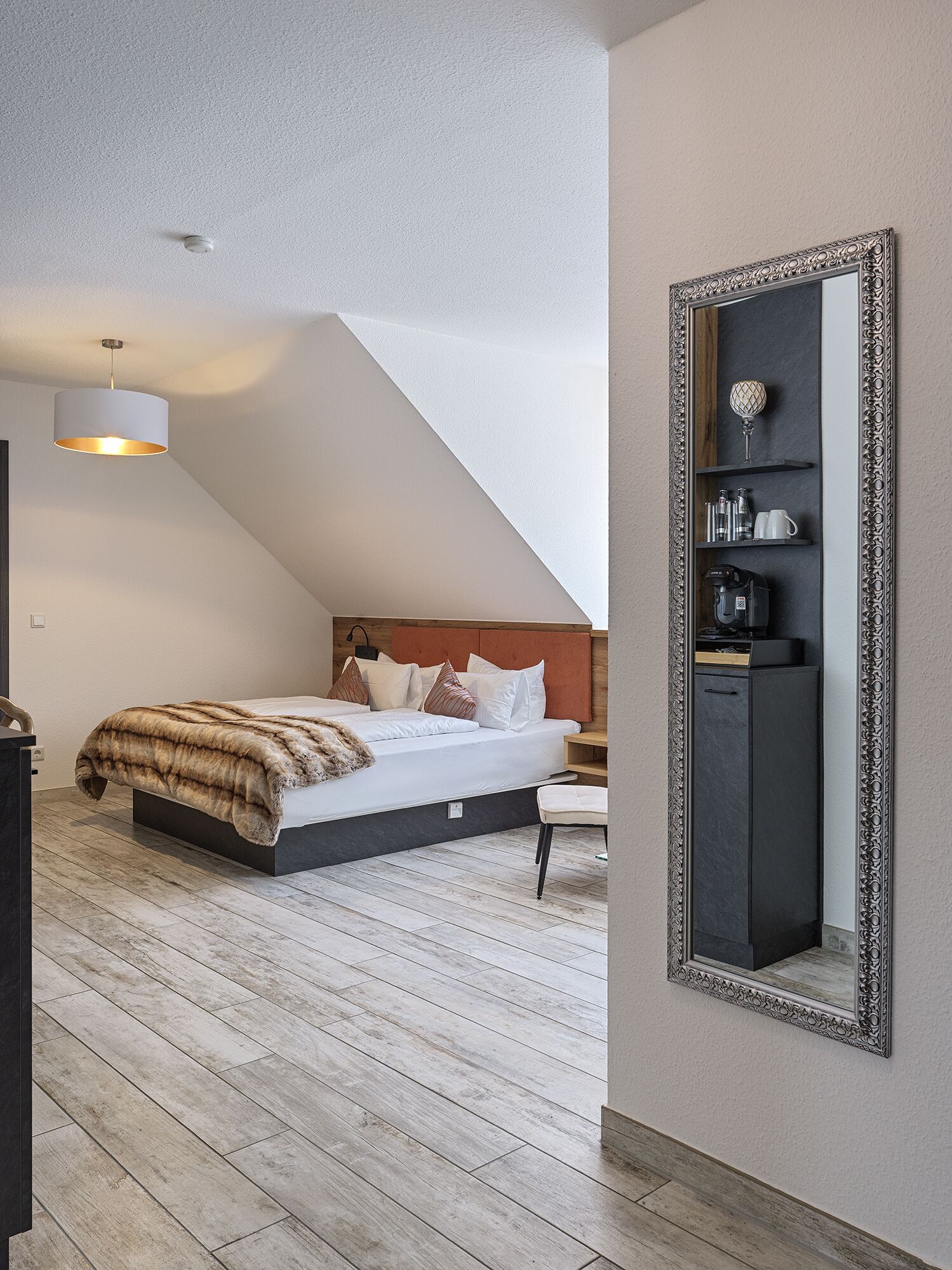 A modern bedroom with a sloped ceiling, a large bed with a fur throw, a bedside table, a dresser, and a mirror with a decorative frame.