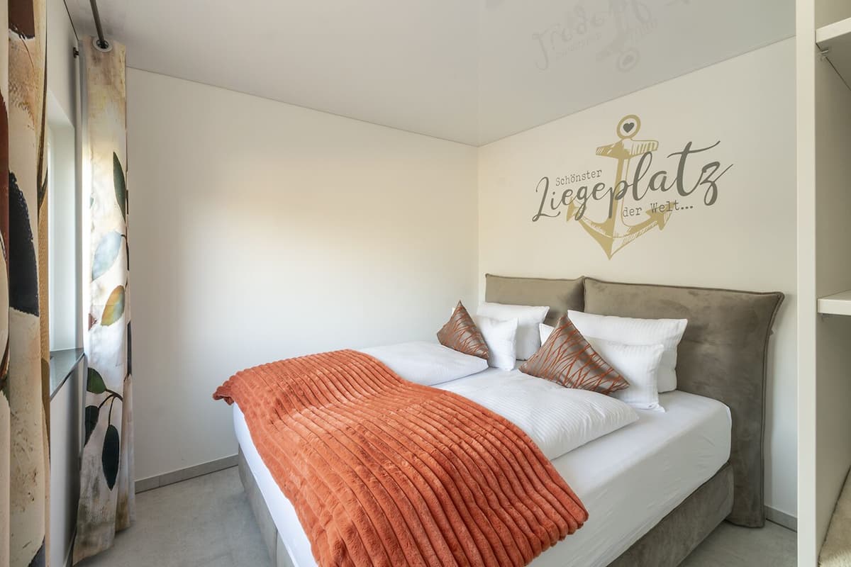 Un lit bien fait, avec une couverture marron et des oreillers blancs, un mur orné d’un panneau décoratif et un rideau à motifs de feuilles.