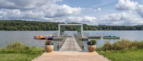 Un muelle de madera que conduce a una estructura con paredes de vidrio junto a un lago con barcos y vegetación.