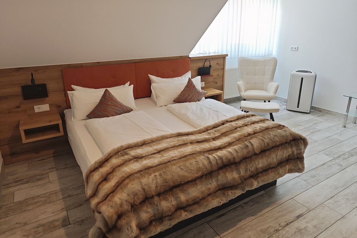Une chambre moderne avec une tête de lit en bois, un grand lit recouvert de draps blancs, un plaid en fausse fourrure, une chaise et un radiateur portatif.