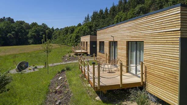 Un hilera de cabañas de madera con terrazas, rodeadas por un campo cubierto de hierba y un bosque denso.