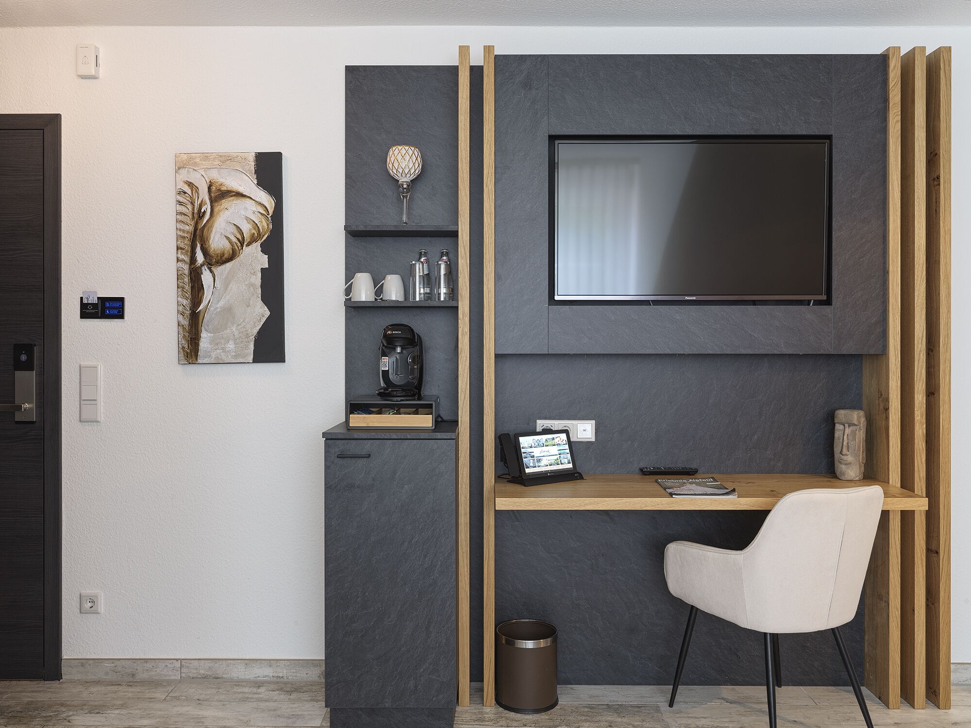 A modern room with a flat-screen TV mounted on a dark wall, a coffee machine, a small shelf with cups and a decorative item, a desk with a chair, and a painting on the wall.
