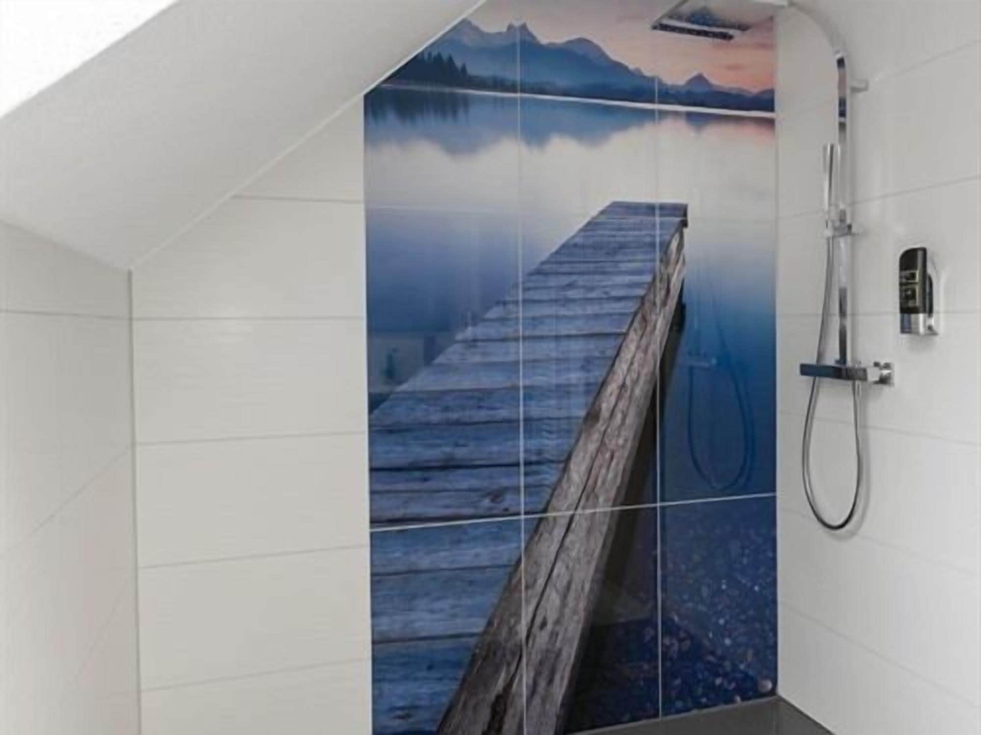 A bathroom with a wooden dock extending into a lake, a shower, and a tiled wall with a mountain and sunset view.