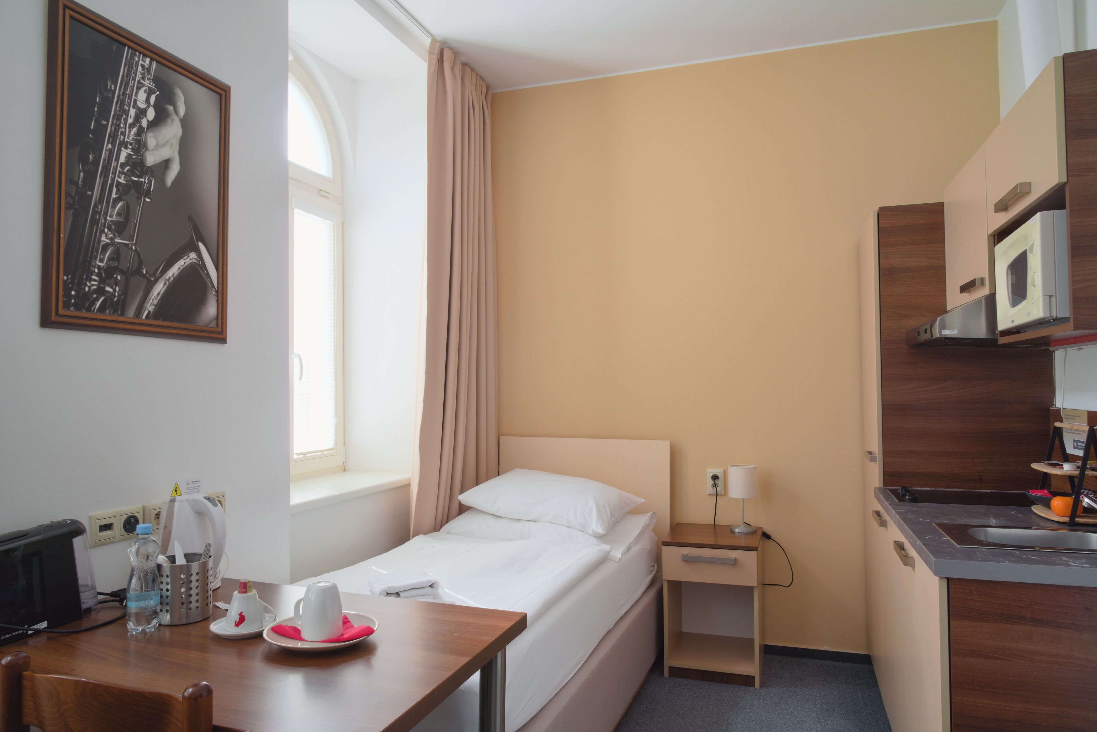 Habitación de hotel con cama, mesita de noche, pequeño escritorio con silla, ventana con cortinas, microondas, lavabo y un cuadro en la pared.