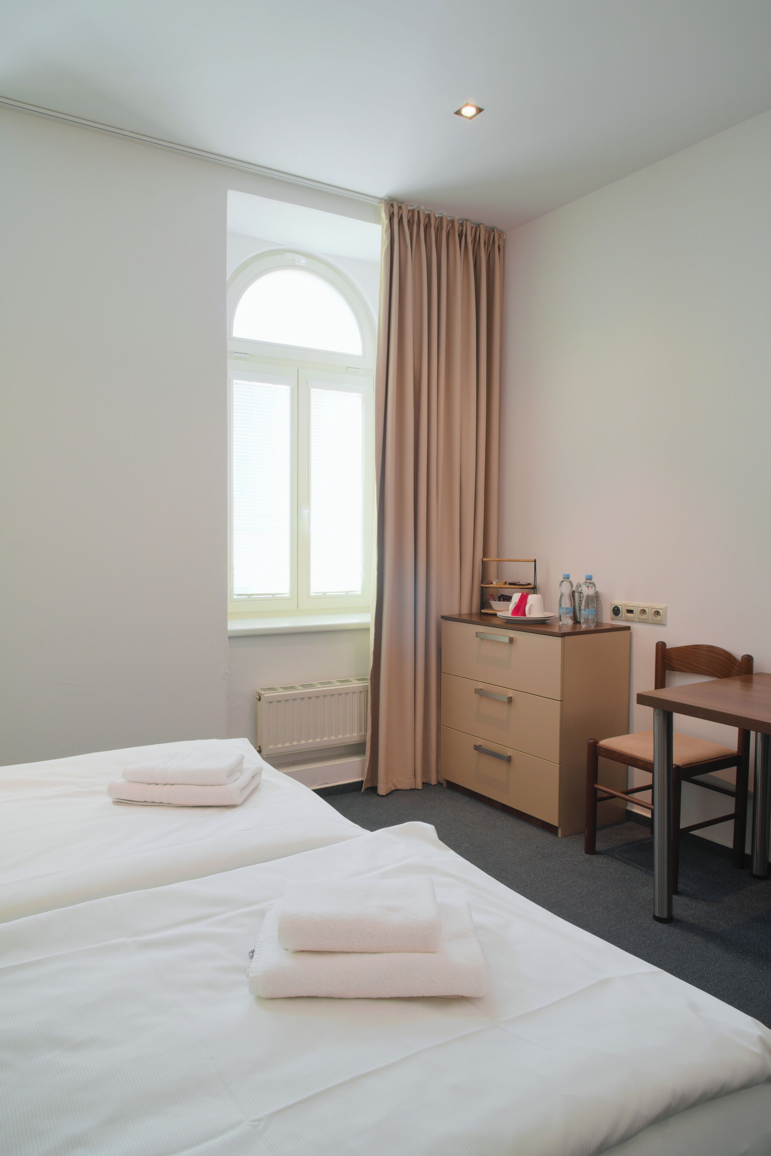 A hotel room with two beds, a desk, a chair, a radiator, and a window with curtains.