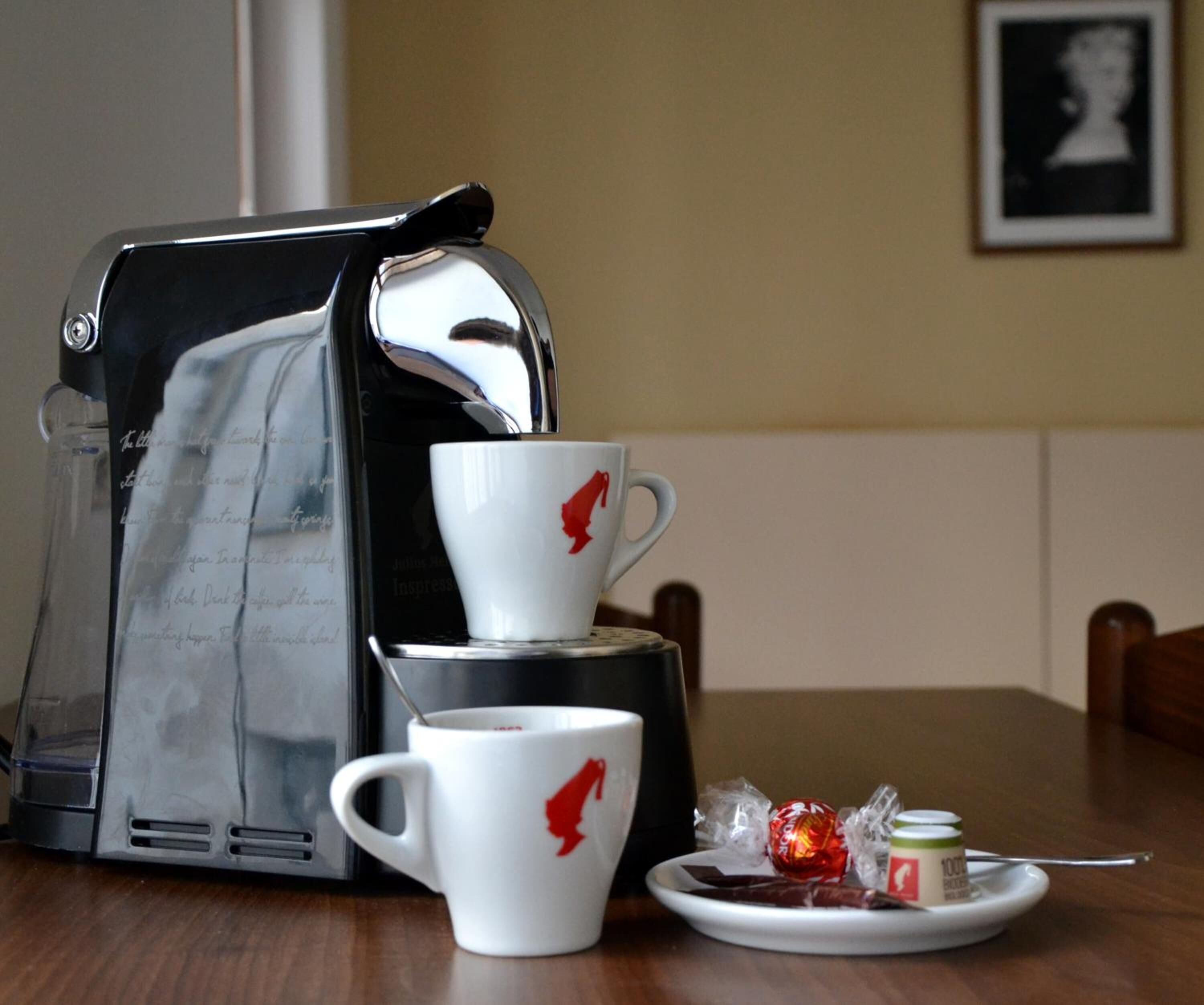 A coffee maker, a white cup with a red logo, a saucer, a plate with a red candy, and a small jar with a spoon on a wooden table.