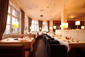 A restaurant with multiple tables and booths, large windows, and hanging lamps. - Hotel Novum (Hinte)