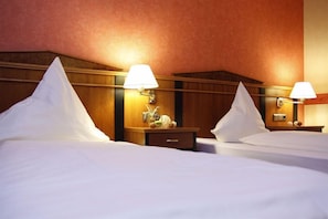 A hotel room with two beds, wooden headboards, bedside tables with lamps, and a small nightstand with a clock and decorative items. - Hotel Novum (Hinte)