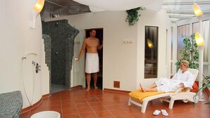 Ett spa-rum med ett kaklat golv, en massagebänk, en person inlindad i en handduk och en person som sitter ned och bär en badrock.