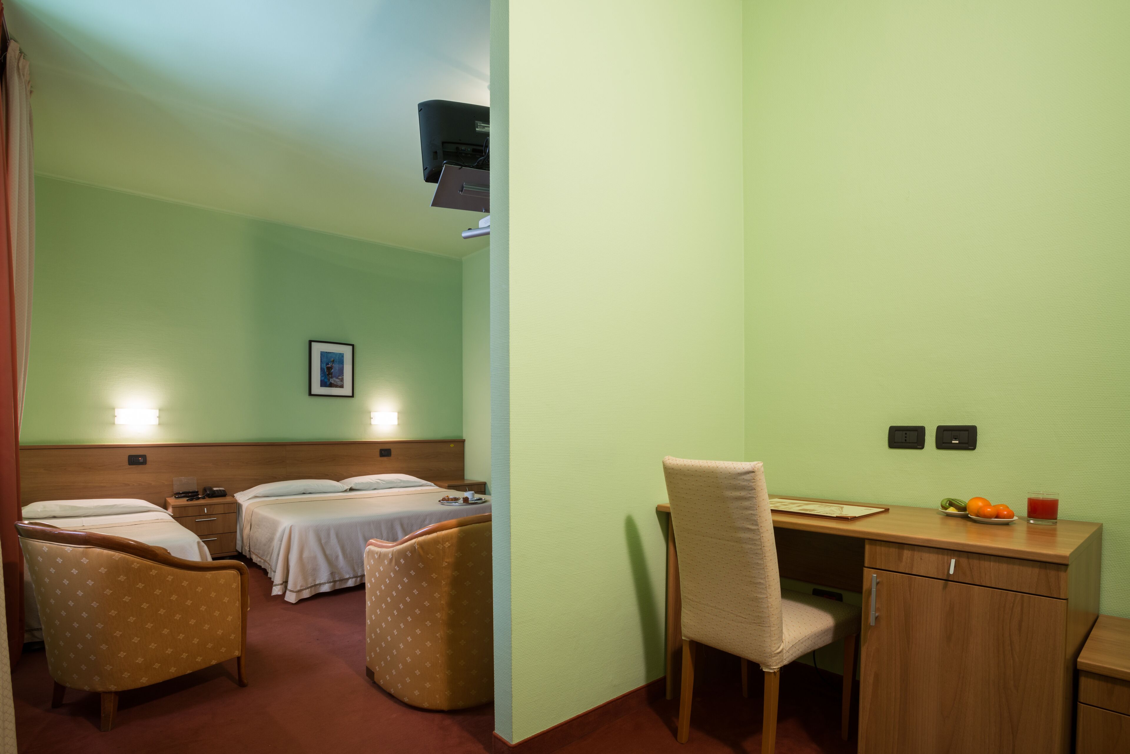 Habitación de hotel con dos camas, un escritorio, una silla y una pequeña mesa con una fuente de fruta y un vaso de jugo.