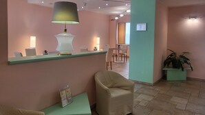 A reception area with a reception desk, chairs, a lamp, and a potted plant. - Hotel Lux (Alessandria)