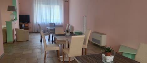 Una sala de estar moderna con comedor, que presenta un amplio ventanal con cortinas translúcidas, un televisor de pantalla plana, una mesa de comedor con sillas y un equipo de aire acondicionado integrado.