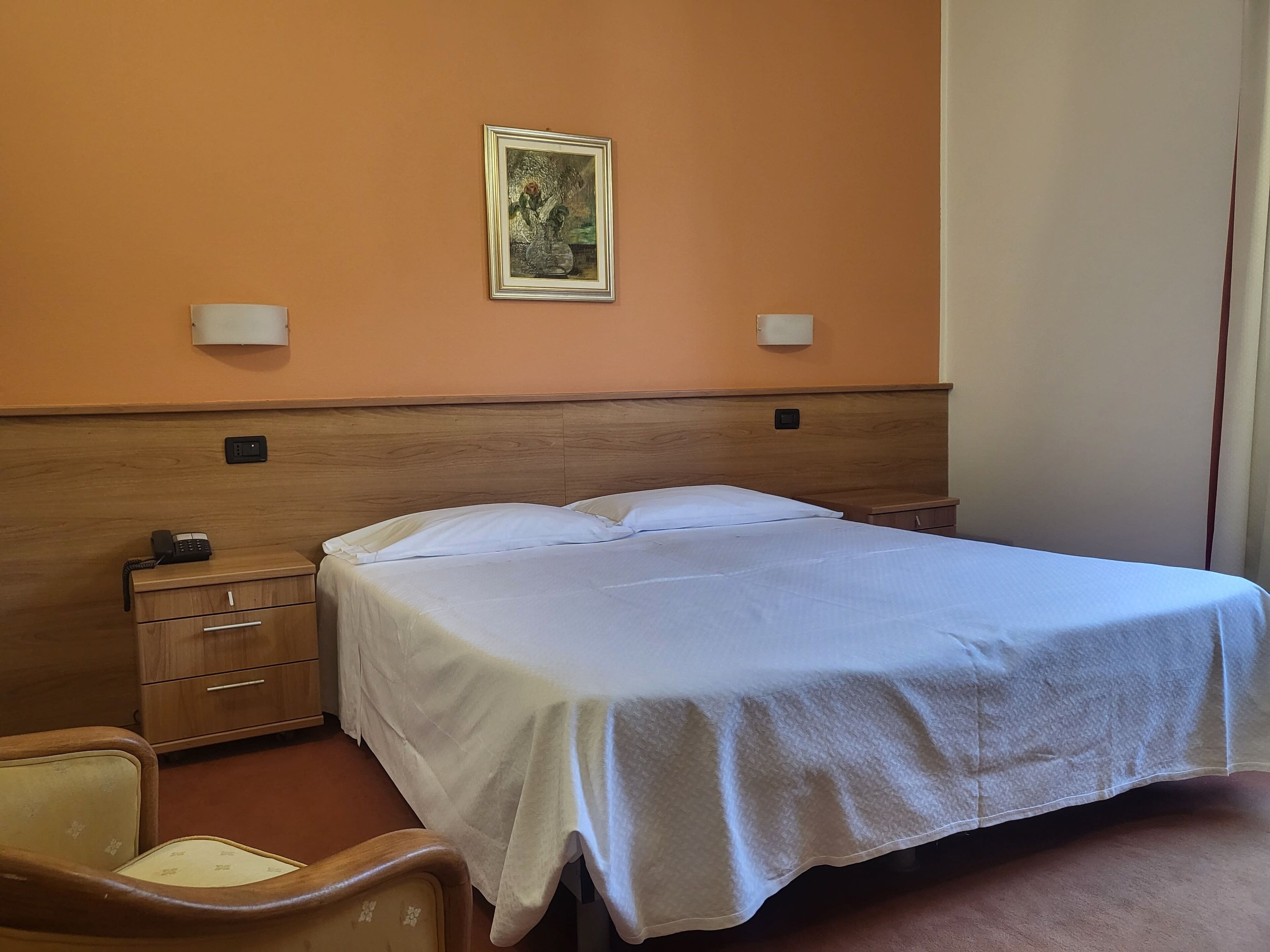 Ein Hotelzimmer mit einem Bett, einem Nachttisch aus Holz mit einem Telefon, einem Sessel und einem Gemälde an der Wand.