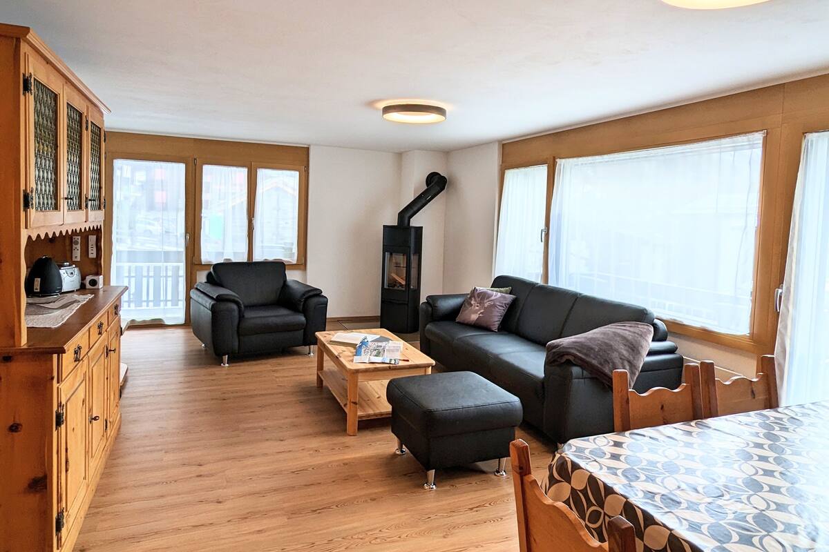 A cozy living room with a sofa, armchair, and ottoman, wooden floor, and a built-in wood stove.